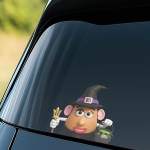 May include: A whimsical car window decal featuring a potato-shaped character dressed as a witch. The character wears a black witch hat with a purple band and holds a golden trident and a cauldron with green liquid. The decal is set against a car window.