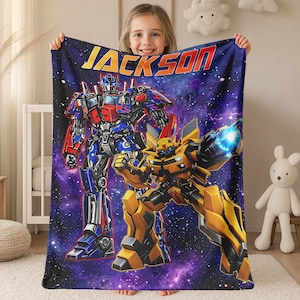 May include: A rectangular fleece blanket featuring a deep purple and blue galaxy background with stars. The blanket displays the name JACKSON in yellow letters at the top, with Optimus Prime and Bumblebee robots printed in the center.