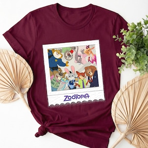 May include: Burgundy t-shirt featuring a framed image of Zootopia characters. The image includes a variety of animated animals in a group photo. The word "Zootopia" is printed in white at the bottom of the frame. The shirt is made of a soft material.