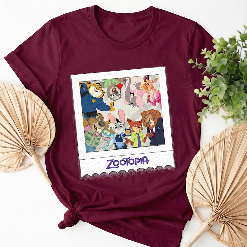 May include: Burgundy t-shirt featuring a framed image of Zootopia characters. The image includes a variety of animated animals in a group photo. The word "Zootopia" is printed in white at the bottom of the frame. The shirt is made of a soft material.