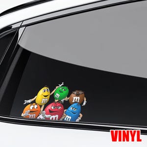 May include: A vinyl decal featuring six colorful M&M'S characters. The characters are in various poses and colors: yellow, green, brown, orange, red, and blue. The word "VINYL" is printed in red at the bottom.