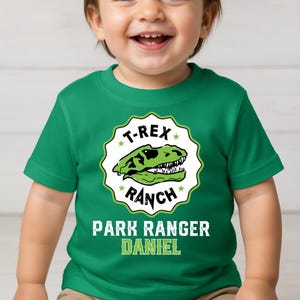 May include: A green t-shirt featuring a dinosaur skull graphic with the text "T-REX RANCH". Below the graphic, the text reads "PARK RANGER DANIEL". The shirt is designed for children.