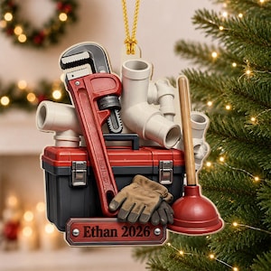 May include: A decorative hanging ornament featuring a red pipe wrench, a black and red toolbox, white PVC pipes, a brown work glove, and a red plunger. A rectangular sign at the bottom displays the text Ethan 2026 in a dark red font.
