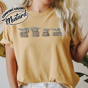 Puede incluir: Una camiseta de manga corta color amarillo mostaza con un estampado gr&aacute;fico de cuatro peque&ntilde;as criaturas verdes con orejas y ojos grandes, cada una con una t&uacute;nica color canela. El texto COMFORT COLORS MUSTARD aparece en una insignia circular.