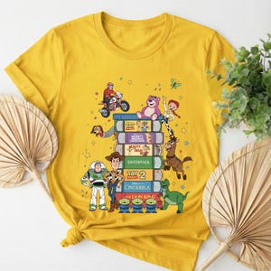 Może przedstawiać: Ż&oacute;łta koszulka z nadrukiem przedstawiającym stos kaset VHS z tytułami film&oacute;w animowanych i postaciami z kresk&oacute;wek. Projekt zawiera postacie z Toy Story, Kopciuszka i Kr&oacute;la Lwa.