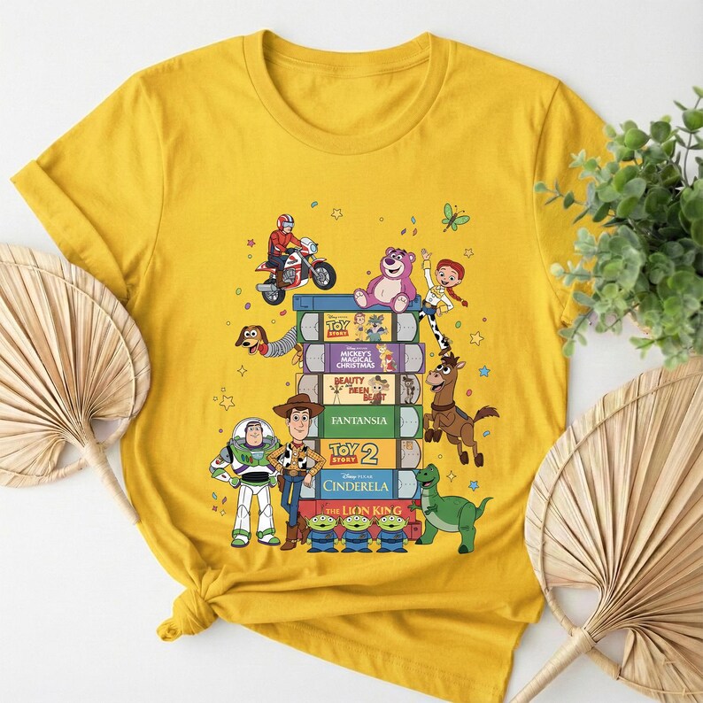 Może przedstawiać: Ż&oacute;łta koszulka z nadrukiem przedstawiającym stos kaset VHS z tytułami film&oacute;w animowanych i postaciami z kresk&oacute;wek. Projekt zawiera postacie z Toy Story, Kopciuszka i Kr&oacute;la Lwa.