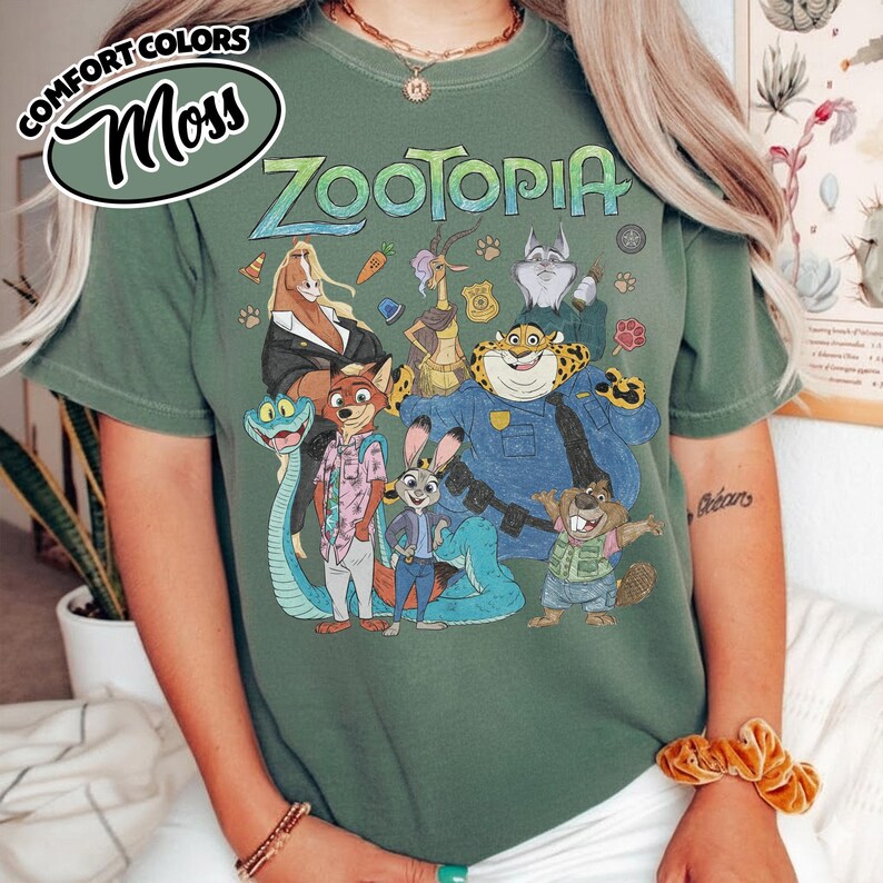 May include: A moss green short-sleeve t-shirt featuring a graphic print of various Zootopia characters including Nick Wilde, Judy Hopps, and Chief Bogo. The text Zootopia is printed in a stylized font above the characters, with a Comfort Colors logo.