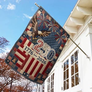 May include: A patriotic flag with a quilted design, featuring red and white stripes, blue sections with fireworks, and an eagle. The flag displays the text "1776-2026" and the number "250". It is mounted on a pole attached to a white building.