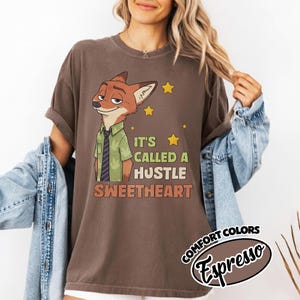 Puede incluir: Una camiseta marr&oacute;n de manga corta con el gr&aacute;fico de un zorro de dibujos animados con camisa verde y corbata. El texto dice IT'S CALLED A HUSTLE SWEETHEART, acompa&ntilde;ado de estrellas amarillas. La camiseta tiene cuello redondo y corte holgado.
