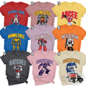 May include: A collection of nine short-sleeved t-shirts in various colors, each featuring a unique Transformers character graphic and their name printed in an arched font above the character, including Optimus Prime, Grimlock, Arcee, Bumblebee, Ironhide, Megatron, Ratchet, Shockwave, and Soundwave.
