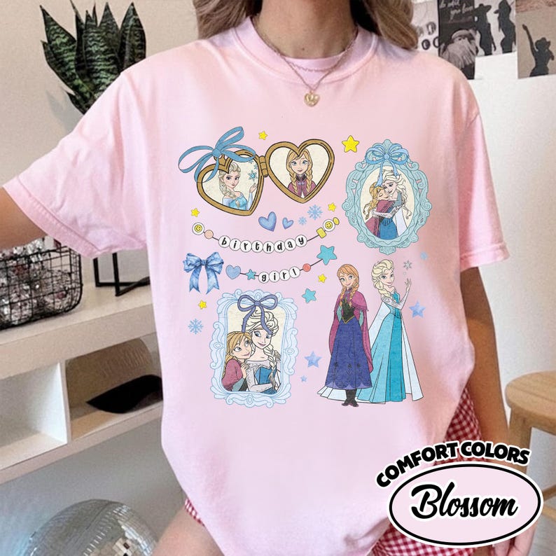 May include: A light pink t-shirt featuring a graphic print of characters from Frozen. The design includes heart-shaped lockets, ornate blue frames, and the text "birthday girl" arranged in a string. Anna and Elsa are depicted in various poses.