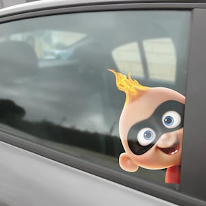 May include: A car window decal featuring the character Jack-Jack Parr from The Incredibles. The decal shows Jack-Jack's face with a black mask, blue eyes, and a flame-like hairstyle. The decal is peeking out of a car window.