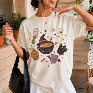 May include: A cream-colored t-shirt featuring a graphic print of various Halloween-themed baking items, including a smiling cauldron with a witch hat, a pumpkin cupcake, a whisk, a piping bag, a bat-winged mug, and a black oven mitt.