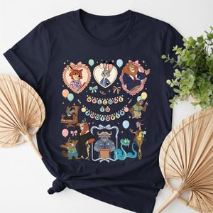K&ouml;nnte beinhalten: Marineblaues T-Shirt mit einem farbenfrohen Grafikdesign von Charakteren aus Zootopia. Das Design zeigt gerahmte Portr&auml;ts von Judy Hopps, Nick Wilde und anderen Charakteren, mit dem Text "Welcome to Zootopia" in der Mitte. Ballons und dekorative Elemente umgeben die Charaktere.