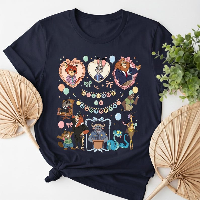 K&ouml;nnte beinhalten: Marineblaues T-Shirt mit einem farbenfrohen Grafikdesign von Charakteren aus Zootopia. Das Design zeigt gerahmte Portr&auml;ts von Judy Hopps, Nick Wilde und anderen Charakteren, mit dem Text "Welcome to Zootopia" in der Mitte. Ballons und dekorative Elemente umgeben die Charaktere.