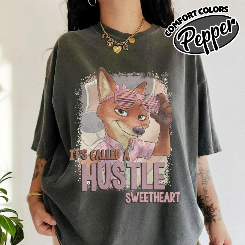 Puede incluir: Una camiseta de manga corta gris carb&oacute;n con el gr&aacute;fico de un zorro de dibujos animados que lleva gafas de sol rosas y una camisa estampada rosa. El texto "IT'S CALLED A HUSTLE SWEETHEART" aparece en letras claras bajo el zorro.