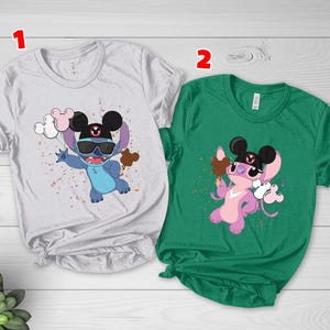 K&ouml;nnte beinhalten: Zwei kurz&auml;rmelige T-Shirts mit Zeichentrickfiguren. Eines ist wei&szlig; mit einer blauen Figur, die eine Sonnenbrille und einen Hut tr&auml;gt und Luftballons und einen Leckerbissen h&auml;lt. Das andere ist gr&uuml;n mit einer rosa Figur, die einen Leckerbissen und Luftballons h&auml;lt.