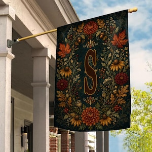 May include: A rectangular garden flag hanging from a gold-colored metal pole. The flag features a dark green background with a central letter S surrounded by a detailed wreath of autumn-themed flowers, orange maple leaves, and small acorns.