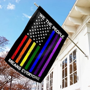 May include: A black flag with a design that combines the American flag with the colors of the rainbow and the text "WE THE PEOPLE MEANS EVERYONE". The flag is hanging from a silver pole on a white house.