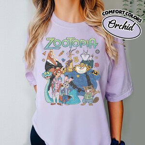 May include: A light purple t-shirt featuring a graphic print of Zootopia characters including Nick Wilde, Judy Hopps, and Chief Bogo. The text Zootopia is printed in a stylized font above the characters. A Comfort Colors Orchid label is visible.