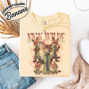 May include: A pale yellow t-shirt featuring a graphic print of the character Nick Wilde in various poses. The text "NICK WILDE" is printed in a retro font above the character illustrations. The shirt has a crew neckline and a Comfort Colors label.