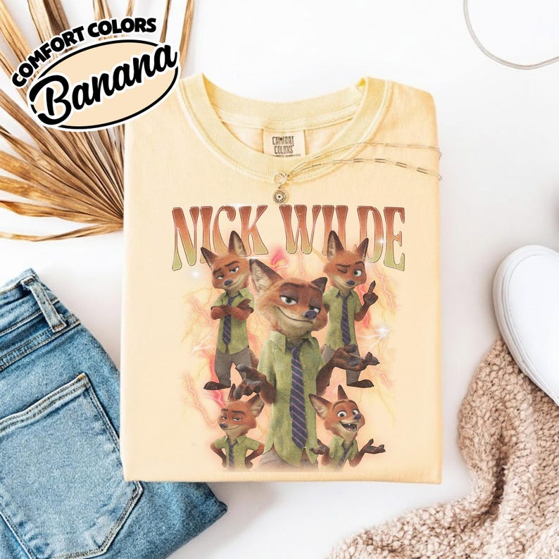 May include: A pale yellow t-shirt featuring a graphic print of the character Nick Wilde in various poses. The text "NICK WILDE" is printed in a retro font above the character illustrations. The shirt has a crew neckline and a Comfort Colors label.
