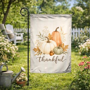 May include: A rectangular garden flag featuring a watercolor illustration of three pumpkins in shades of orange, white, and sage green, surrounded by autumn leaves, wheat, and an acorn. The word thankful is printed in brown cursive script below.