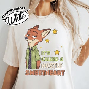 Puede incluir: Una camiseta color crema con el gr&aacute;fico de un zorro de dibujos animados que viste una camisa verde con estampado de hojas y una corbata oscura. El texto dice IT'S CALLED A HUSTLE SWEETHEART, junto a estrellas amarillas. Logo COMFORT COLORS White.