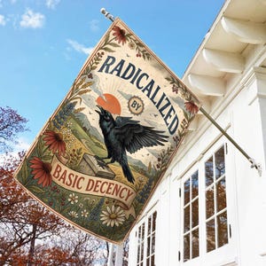 May include: A rectangular flag with the words "RADICALIZED BY BASIC DECENCY" and a detailed illustration of a black crow, a sunset, and a landscape. The flag is attached to a pole and hangs outside a white building.