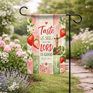 May include: A garden flag with a floral and strawberry design. The flag features the text "Taste & See - That The Lord Is Good" with a cross and strawberries. The background has a pink and green color scheme.