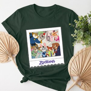 May include: Dark green t-shirt featuring a framed image of Zootopia characters. The image includes Judy Hopps, Nick Wilde, and other characters from the animated film. The word "Zootopia" is printed in blue at the bottom of the frame.