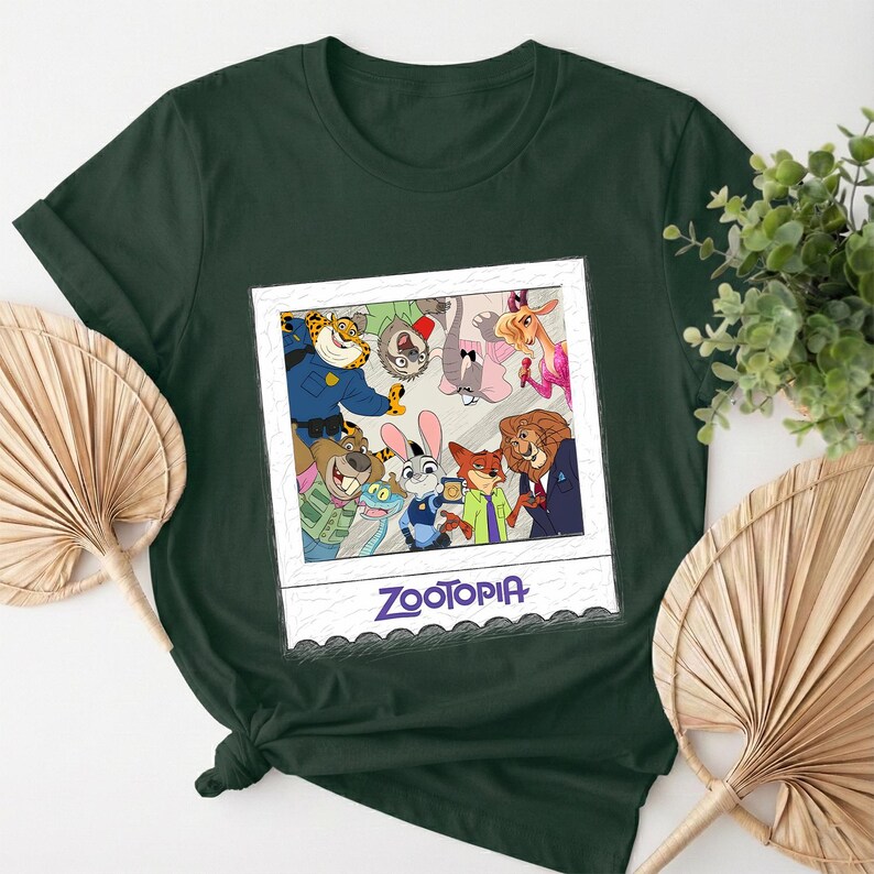 May include: Dark green t-shirt featuring a framed image of Zootopia characters. The image includes Judy Hopps, Nick Wilde, and other characters from the animated film. The word "Zootopia" is printed in blue at the bottom of the frame.
