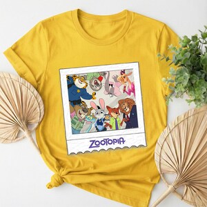 May include: Mustard yellow t-shirt featuring a framed image of Zootopia characters. The image includes Judy Hopps, Nick Wilde, and other characters from the animated film. The word "Zootopia" is printed in a purple font at the bottom of the image.