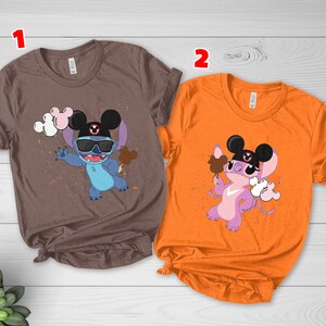 K&ouml;nnte beinhalten: Zwei T-Shirts, eines braun und eines orangefarben, mit einer Cartoonfigur, die eine Sonnenbrille und einen Mickey Mouse-Hut tr&auml;gt. Die Figur h&auml;lt ein Eis und Luftballons. Die T-Shirts sind kurz&auml;rmlig.