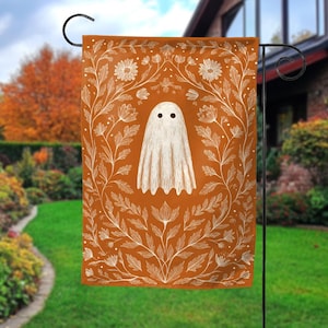 May include: A Halloween garden flag featuring a white ghost with black eyes on an orange background. The ghost is surrounded by a white floral design. The flag is hanging on a black metal stand in a garden setting.