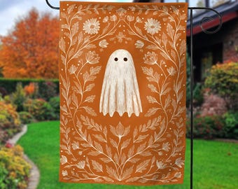 May include: A Halloween garden flag featuring a white ghost with black eyes on an orange background. The ghost is surrounded by a white floral design. The flag is hanging on a black metal stand in a garden setting.
