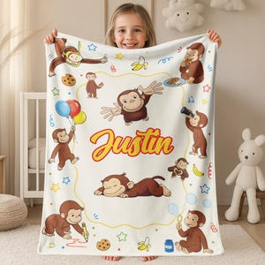 May include: A white fleece blanket featuring a repeating pattern of Curious George monkeys in various poses, including eating cookies, holding balloons, and drawing. The name Justin is printed in large, bold, orange and yellow script in the center.