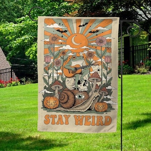 May include: A beige garden flag with a retro design featuring a frog playing guitar atop a skull and snail. The flag includes a sun, moon, flowers, bats, and the text "STAY WEIRD" in orange.