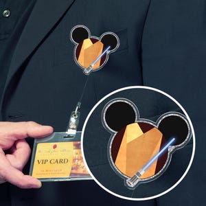 May include: A clear badge reel featuring a Mickey Mouse-shaped design with a brown geometric shape and a blue lightsaber. The badge reel is attached to a VIP card holder.
