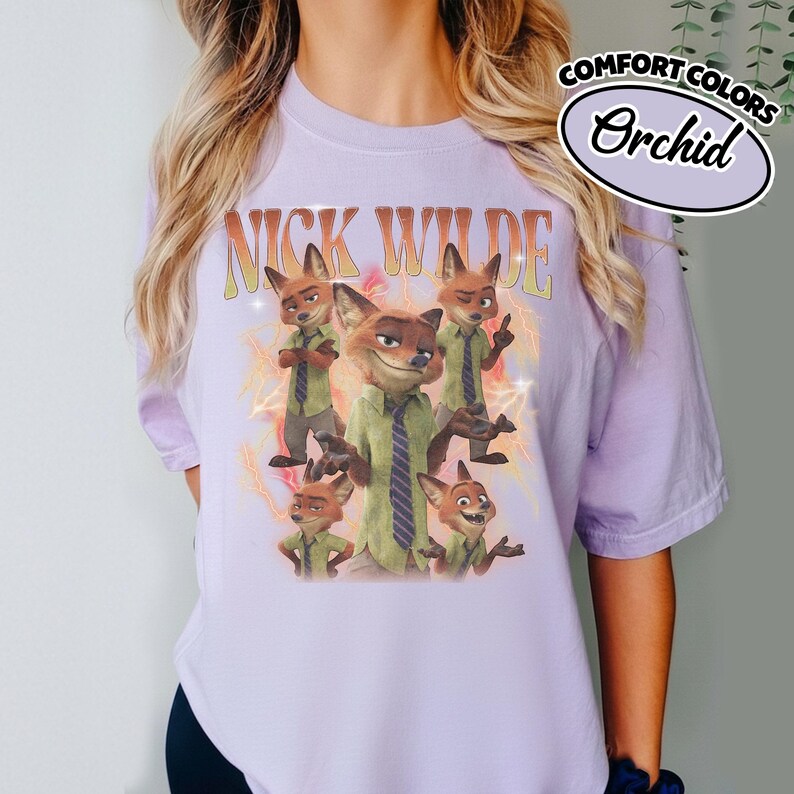 May include: A light purple t-shirt featuring a graphic print of the character Nick Wilde in various poses. The text "NICK WILDE" appears in a curved orange font above the character. The design includes yellow lightning bolt accents.