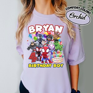 May include: A light purple t-shirt featuring a graphic print with the text BRYAN, 4, and BIRTHDAY BOY. The design includes cartoon superhero characters, colorful balloons, and a birthday cake with candles. A label reads COMFORT COLORS Orchid.