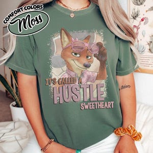 Puede incluir: Una camiseta verde musgo con un gr&aacute;fico de un zorro de dibujos animados con gafas de sol de persiana rosas. El zorro lleva una camisa de cuello rosa claro. Debajo del zorro, el texto dice IT'S CALLED A HUSTLE SWEETHEART en letras oscuras.