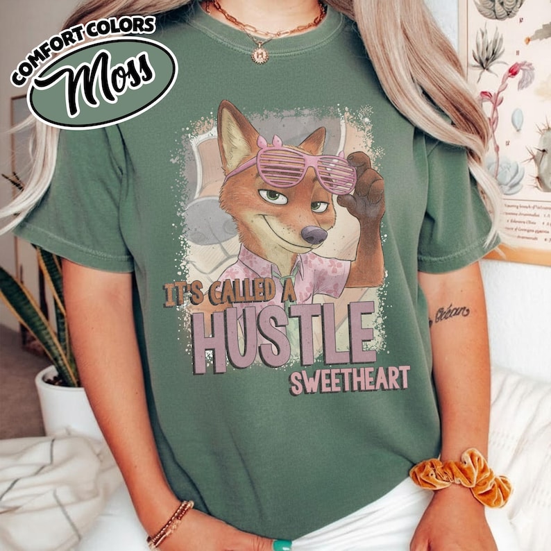 Puede incluir: Una camiseta verde musgo con un gr&aacute;fico de un zorro de dibujos animados con gafas de sol de persiana rosas. El zorro lleva una camisa de cuello rosa claro. Debajo del zorro, el texto dice IT'S CALLED A HUSTLE SWEETHEART en letras oscuras.