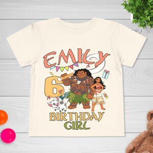 May include: A cream-colored short-sleeve t-shirt featuring a graphic print with the name EMILY, the number 6, and the text BIRTHDAY GIRL. The design includes cartoon characters Maui, Moana, and Pua, along with a gift box, candy, and bunting.