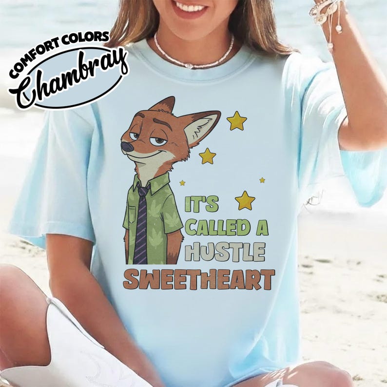 Puede incluir: Una camiseta azul claro con el gr&aacute;fico de un zorro de dibujos animados que lleva una camisa verde y una corbata oscura. El texto dice IT'S CALLED A HUSTLE SWEETHEART, junto a iconos de estrellas amarillas. El logo de Comfort Colors est&aacute; arriba.