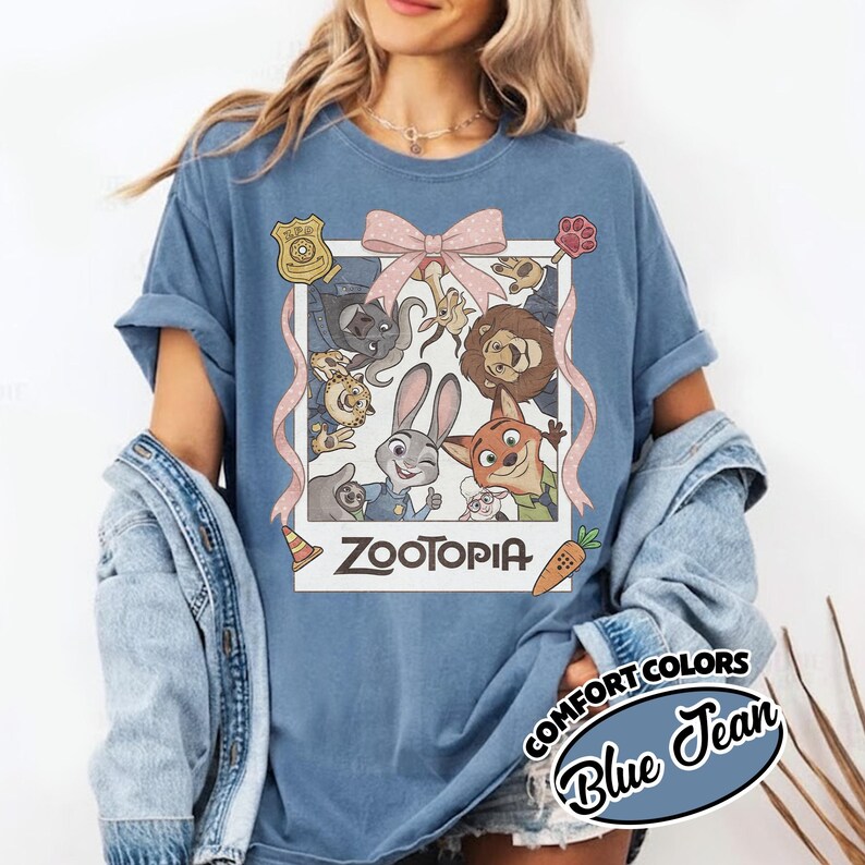 May include: A blue jean colored t-shirt featuring a graphic print of Zootopia characters inside a white polaroid frame. The design includes a pink bow, a police badge, a paw print, a traffic cone, and a carrot. The text Zootopia is printed below.