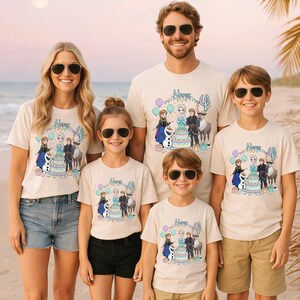 May include: A group of five people wearing matching cream-coloured t-shirts with a colourful graphic featuring characters, a cake, and balloons. The shirts are paired with shorts and sunglasses, posing on a beach.