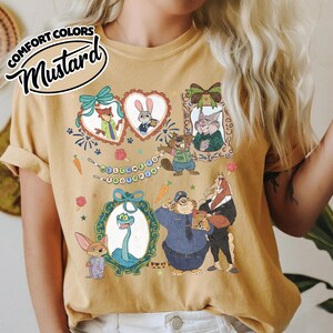 May include: A mustard yellow t-shirt featuring a graphic print of Zootopia characters including Nick Wilde, Judy Hopps, and others in decorative frames. The design includes a banner with the text Welcome to Zootopia and various small icons.