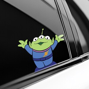 May include: A green alien character sticker with three eyes, wearing a blue uniform with a yellow and orange emblem. The sticker is applied to a car window, with the alien's arms outstretched.