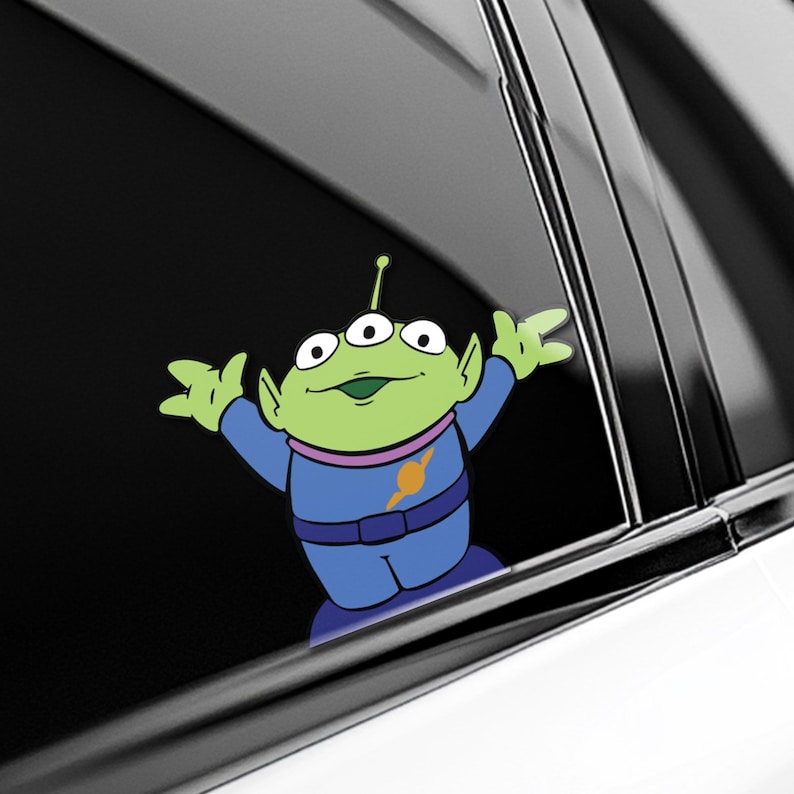 May include: A green alien character sticker with three eyes, wearing a blue uniform with a yellow and orange emblem. The sticker is applied to a car window, with the alien's arms outstretched.